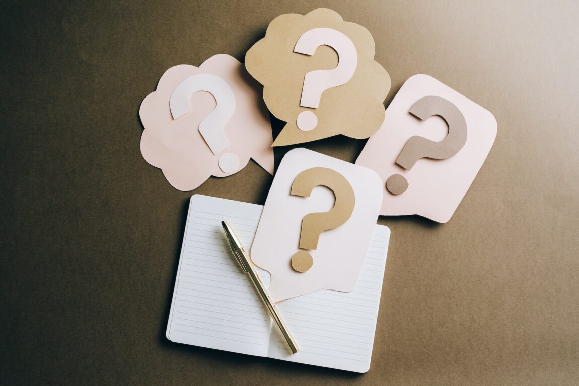 Question marks made of cut out paper laying on a desktop with a notebook open nearby.- Photo by Leeloo Thefirst: https://www.pexels.com/photo/question-marks-on-craft-paper-5428830/