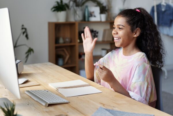 teen girl sitting at her desk, smiling and waving at the monitor Photo by Julia M Cameron: https://www.pexels.com/photo/woman-in-pink-shirt-sitting-by-the-table-while-smiling-4143791/